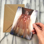 Ballerina Greeting Card, view of a pink organza dress with ribbons and bow against a glowing background, shown with envelope.  Posed with hand for scale.