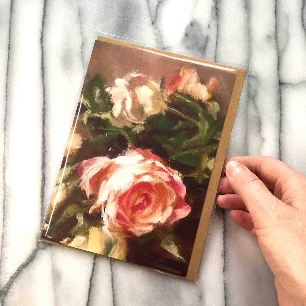 Bouquet of Roses Greeting Card features a bouquet of classic peachy tinted roses clustered with a single red bloom resting near a pair of candlesticks.  Card front, 7"x5" featured with Kraft envelope inside a protective plastic sleeve.  Shown with hand to demonstrate size..