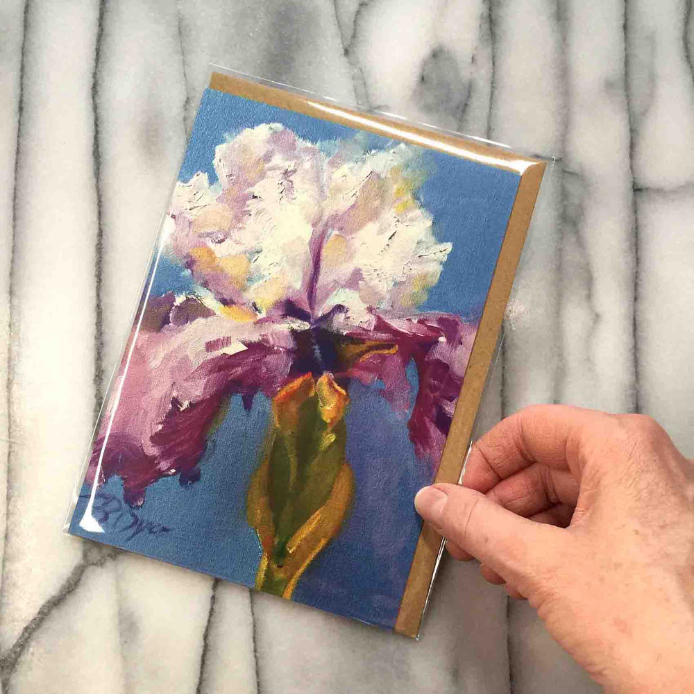
                  
                    Load image into Gallery viewer, Little Iris Greeting Card features a freshly hued single white &amp;amp; purple iris with green stem and is juxtaposed against a vivid blue ground.  Card front, 7&amp;quot;x5&amp;quot; featured with hand to demonstrate card size.
                  
                