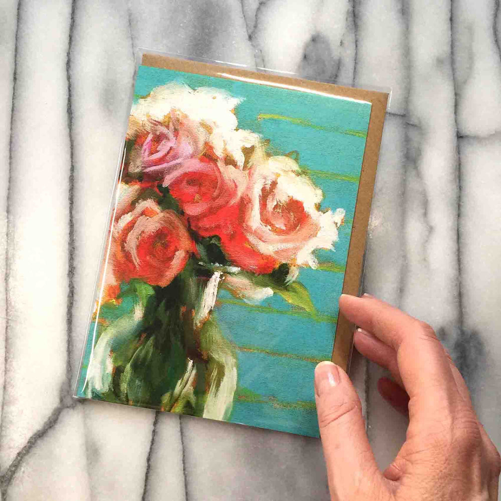 Peonies and Roses Greeting Card features a luscious floral bouquet of red, pink and coral flowers accented by intense green foliage. The modern turquoise and lime background reflects in a glass vase, 7" x 5".  Card front shown with envelope inside protective plastic sleeve, with hand view for size