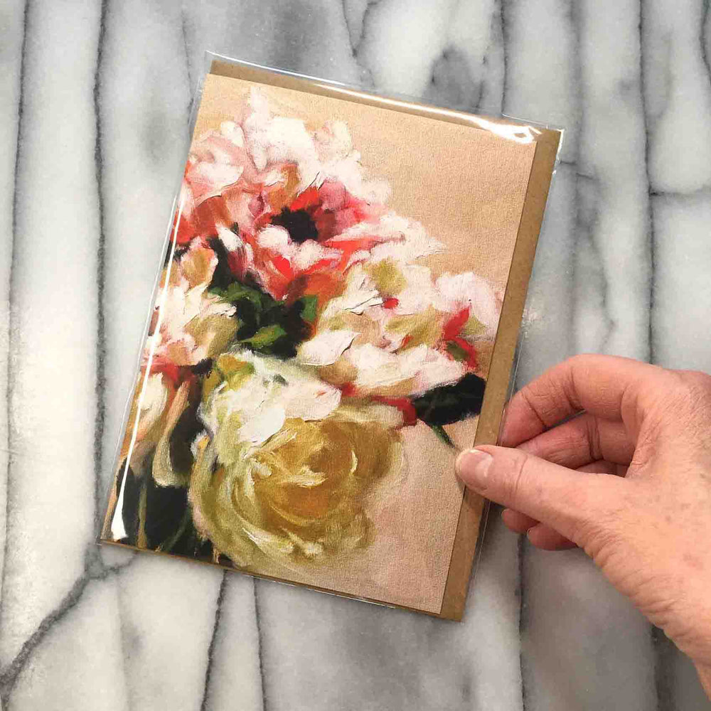 Poppies and Peonies Greeting Card presents an elegant and dreamy floral bouquet of pastel pinks, coral, cream and yellow with leafy accents that rests against a light peach background.   7" x 5”, front view presented with Kraft envelope, inside protective plastic sleeve. Shown with hand to demonstrate card size.